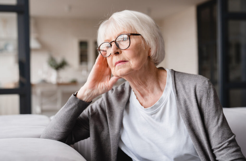 A senior woman with glasses on sitting on a couch and looking out the window with a serious expression.