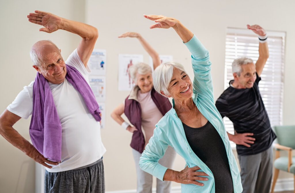 A group of older adults in an exercise class smiling and stretching with one hand over their heads.