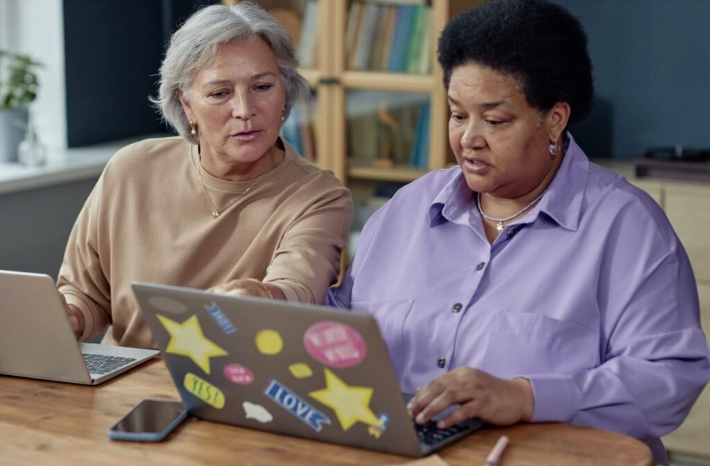 Two seniors on laptops help each other figure out apps they can use to simplify their instrumental daily living activities.