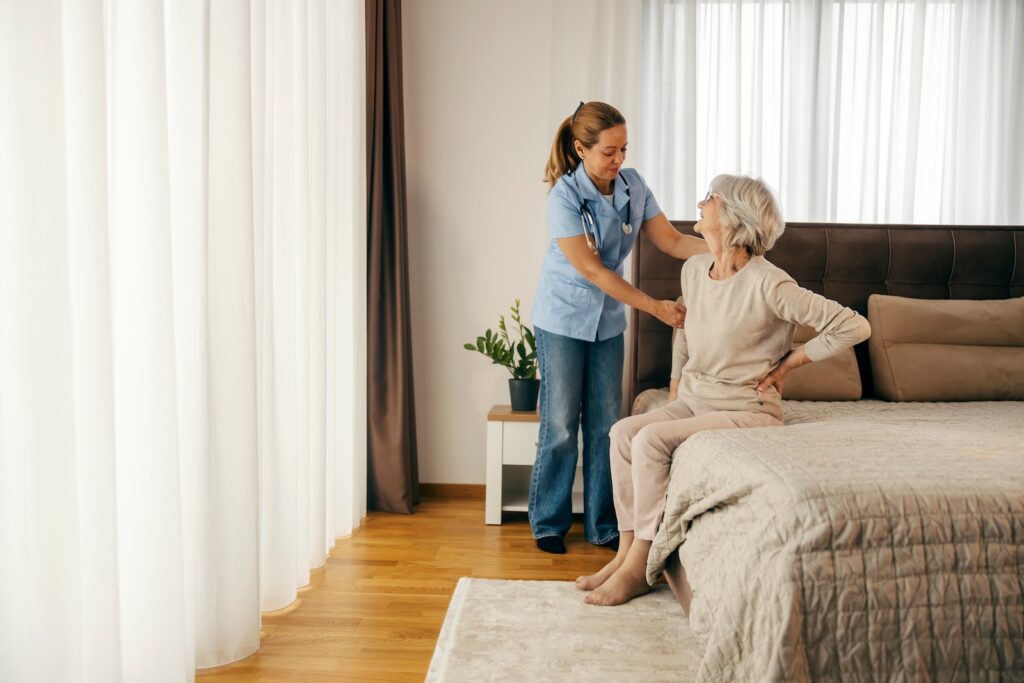 A caregiver assisting an older adult seated on the edge of a bed in a bright, cozy room.