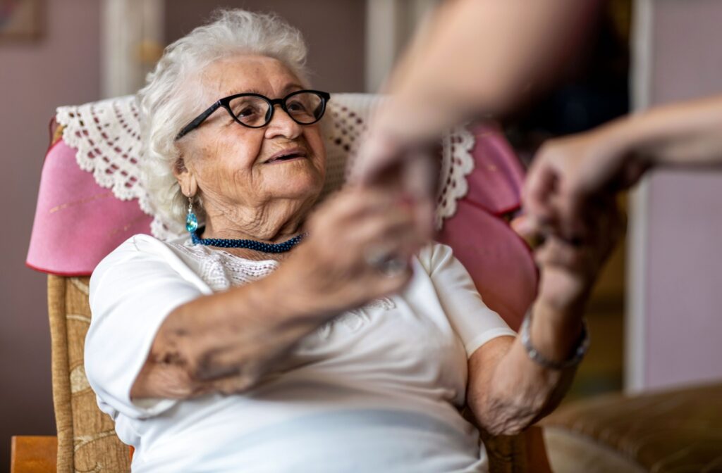 A senior with dementia smiles as their caregiver takes their hands to help them out of their chair.