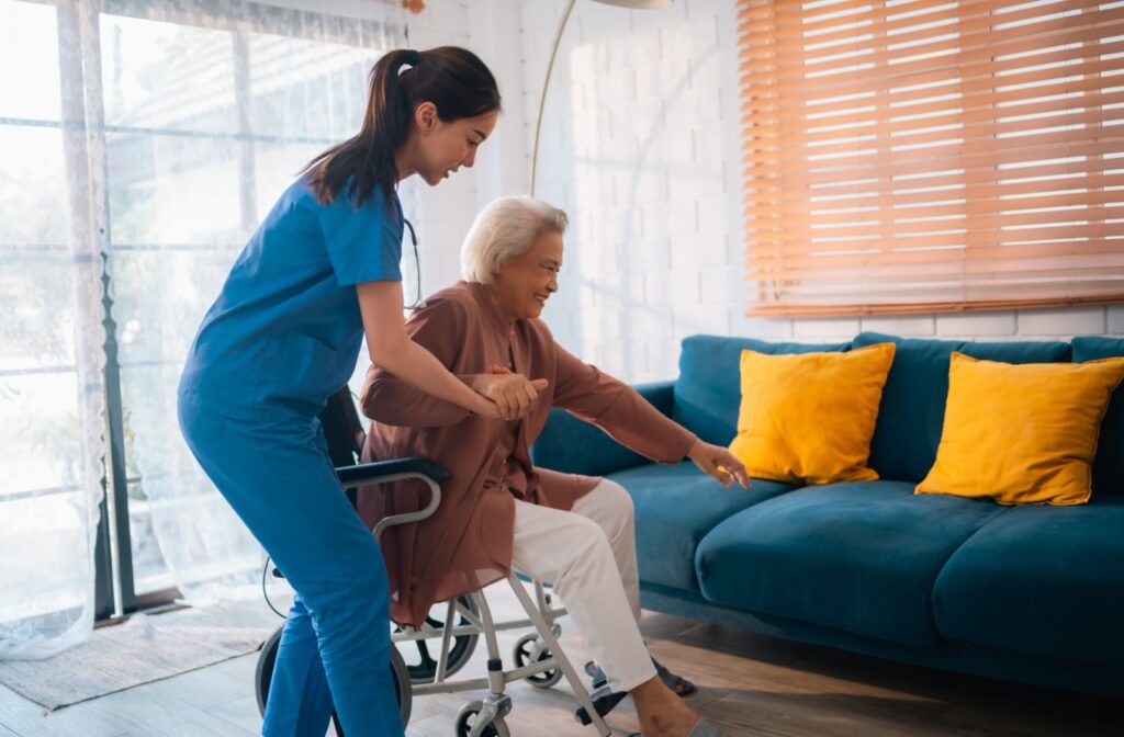 A caregiver assisting an elderly woman out of a wheelchair in a bright living room.
