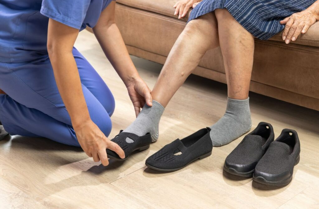 Caregiver kneeling on the floor helping an older adult on a couch put on black slip-resistant slippers.
