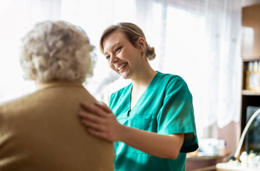 A senior living caregiver in green scrubs smiles while placing a hand on a senior’s back, providing emotional support
