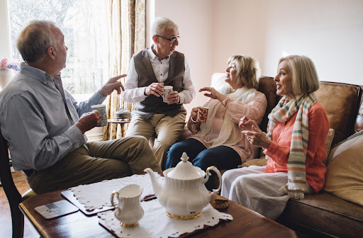 A group of four seniors socialize, two sitting on a couch, and two sitting on chairs close by in a semi-circle. They are holding tea cups.