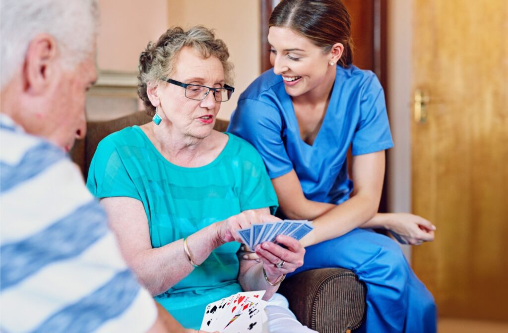 A caregiver smiling and engaging with an elderly woman playing cards during a group activity.