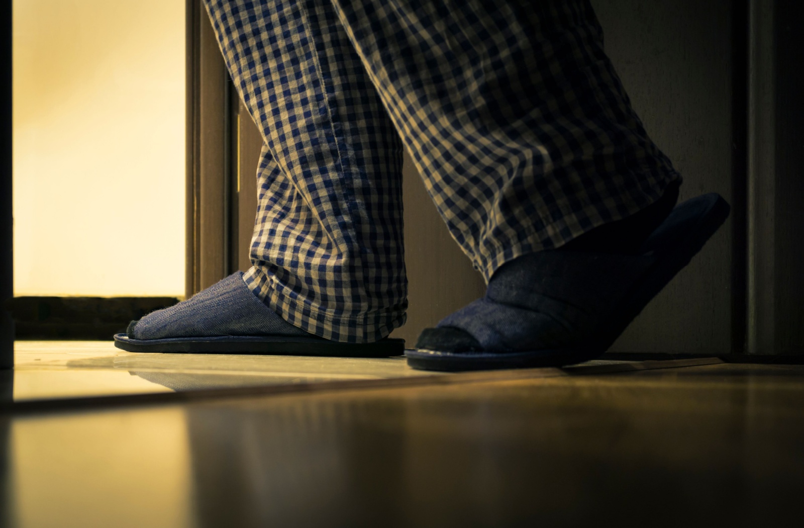 An older adult's legs in pajamas and slippers, walking towards bright light from a dark room.