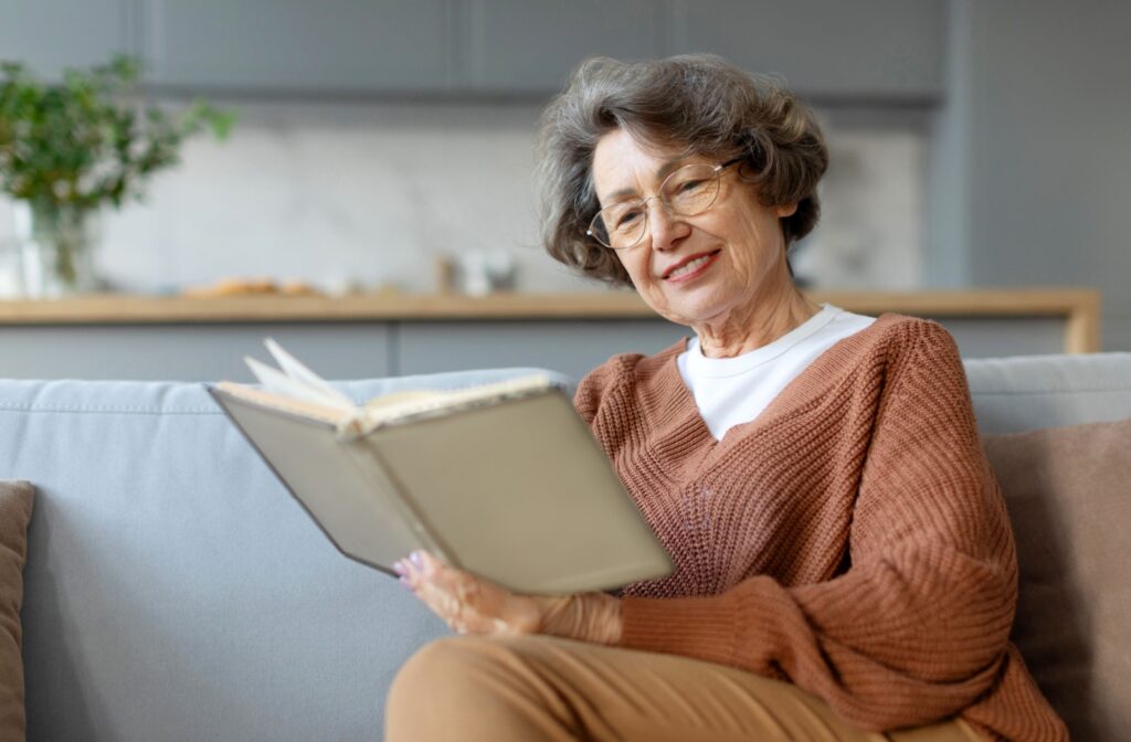 A smiling older adult sitting on their couch and reading a hardcover book.