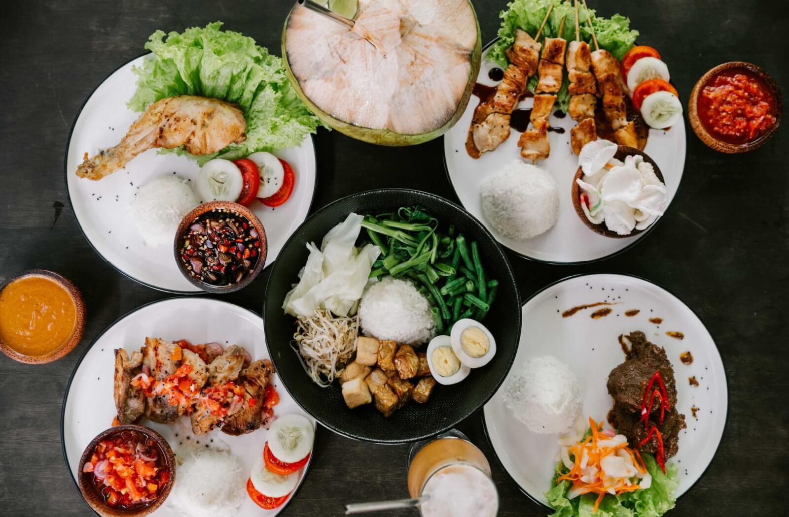 Five plates of Mediterranean meals, full of leafy vegetables and lean meat, sit on a dark table, accented with drinks