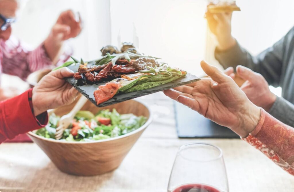 A group of seniors pass plates of food to each other over a shared mealtime with fresh green vegetables