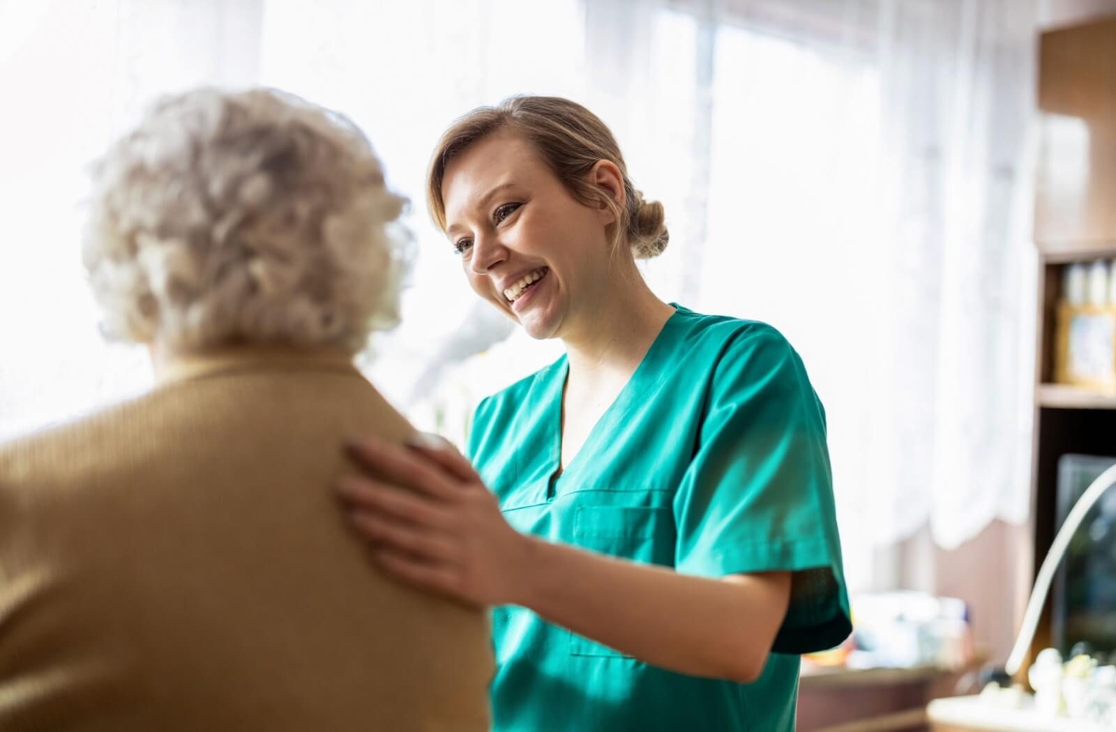 A senior living caregiver in green scrubs smiles while placing a hand on a senior’s back, providing emotional support