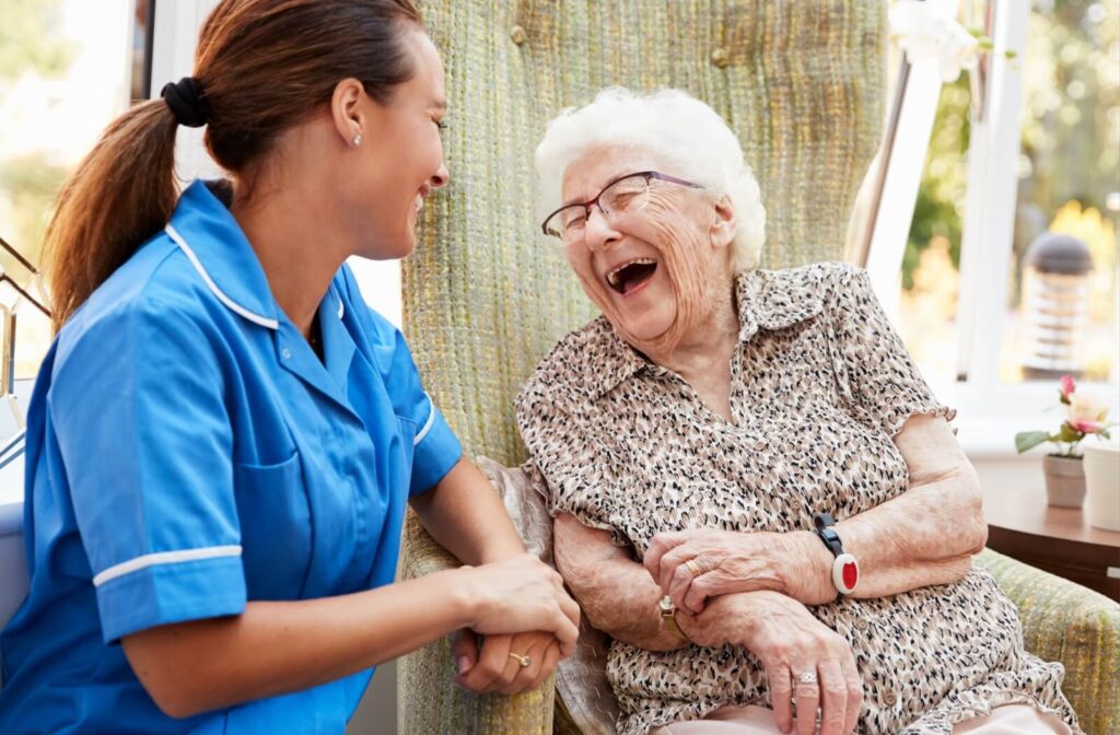 A senior in assisted living reacts with a deep laugh while in conversation with a supportive nurse in blue scrubs